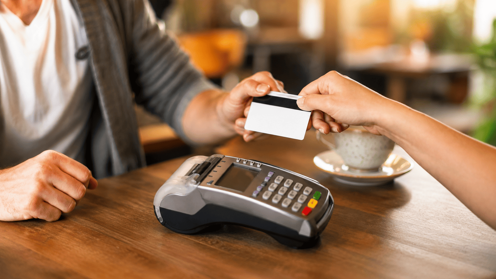 Person swipes a card or hands a card to a cashier at a card payment terminal on a wooden table in a cafe.